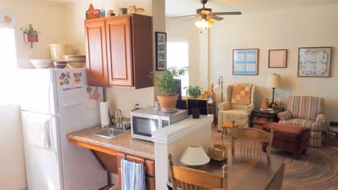 Open-plan interior showing a small kitchen with a fridge, sink, microwave and cabinets opening into a cozy living area with armchairs, a dining table and ceiling fan.
