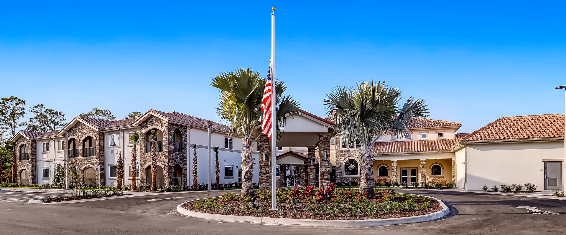 Front exterior of a two-story senior living facility with a circular driveway, flagpole, palm trees, and stone accents under a clear blue sky.