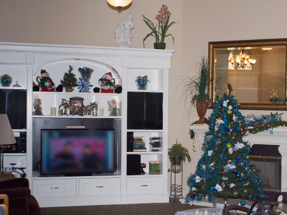 A cozy living room decorated for the holidays with a Christmas tree adorned with blue, green, and purple ornaments and ribbons. The room features a white entertainment center with shelves holding holiday figurines, a small Christmas tree, and decorative items. A flat-screen TV is placed in the center of the entertainment unit. To the right, there is a fireplace with a large mirror above it and a potted plant on the mantel. A chandelier is reflected in the mirror, and a lamp and armchair are partially visible on the left side.
