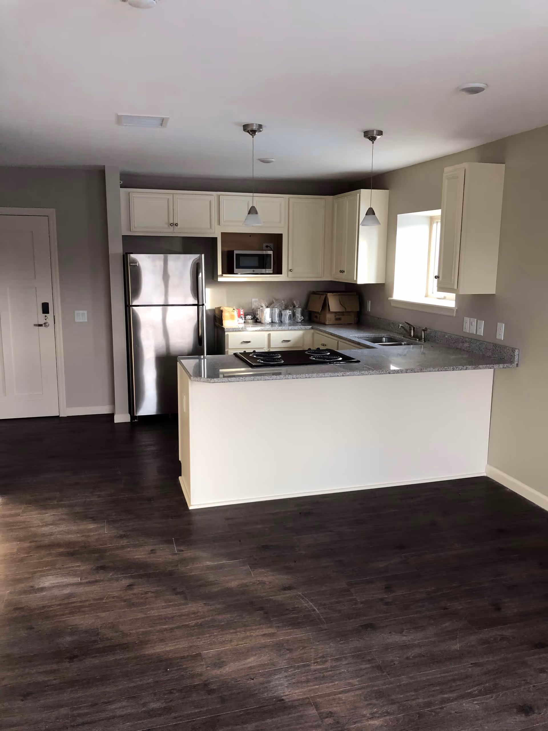 Modern kitchen with white cabinets, stainless steel refrigerator, built-in microwave, and a kitchen island with a cooktop. The kitchen has dark wood flooring and two pendant lights hanging from the ceiling. There is a window above the sink allowing natural light into the space.