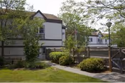 Exterior view of a two-story white nursing facility with brown trim, a gated courtyard, walkway, lawn, trees, and an American flag.