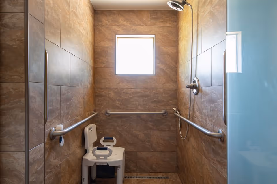 A walk-in shower with brown tiled walls, a small frosted window, a handheld showerhead, and multiple stainless steel grab bars. There is a white shower chair with armrests positioned inside the shower.