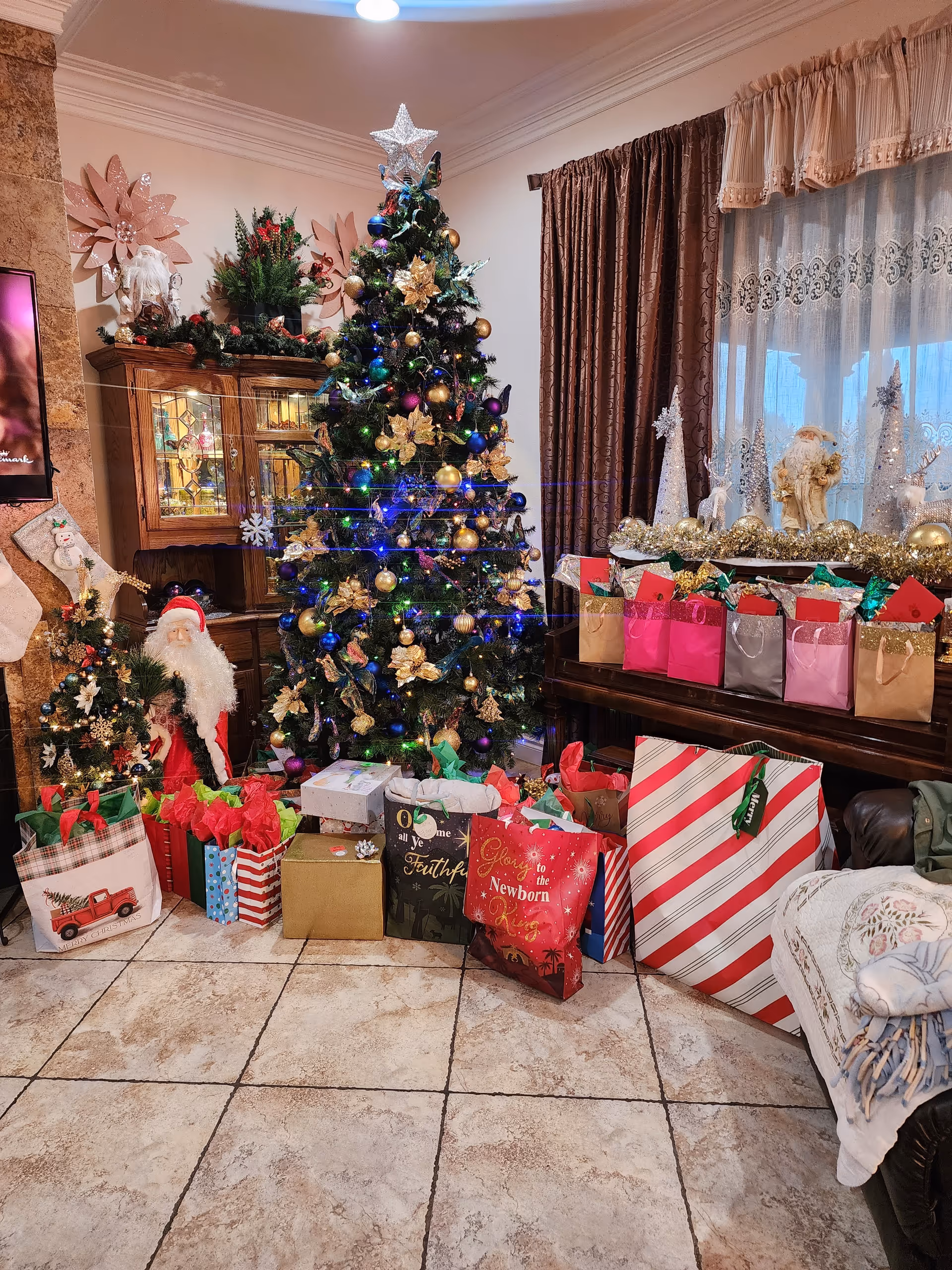 A decorated living room with a Christmas tree adorned with gold and blue ornaments and lights. Numerous wrapped Christmas presents are placed under and around the tree. To the left, there is a small decorated tree and a Santa Claus figure. Behind the tree, a wooden cabinet with glass doors displays various items. On the right side, a table holds more wrapped gifts and festive decorations including a Santa figurine and glittery cone-shaped ornaments. The room has tiled flooring, brown curtains, and lace window coverings.