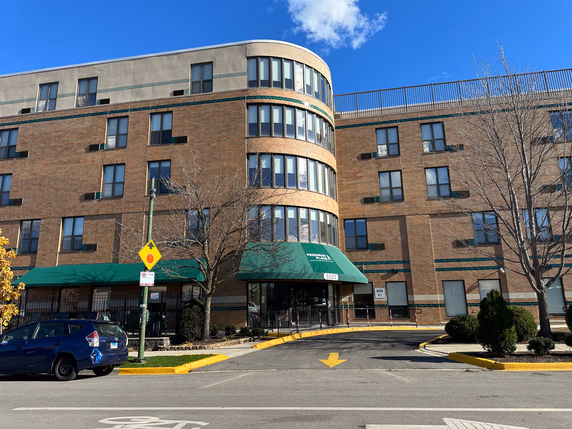 Brick multi-story senior living building with a rounded glass bay and a green awning over the main entrance reading "Friedman Place".