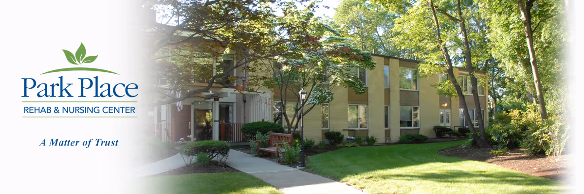 Exterior view of Park Place Rehab & Nursing Center showing a well-maintained garden with green grass, trees, shrubs, a sidewalk, and benches in front of a multi-story building with large windows.
