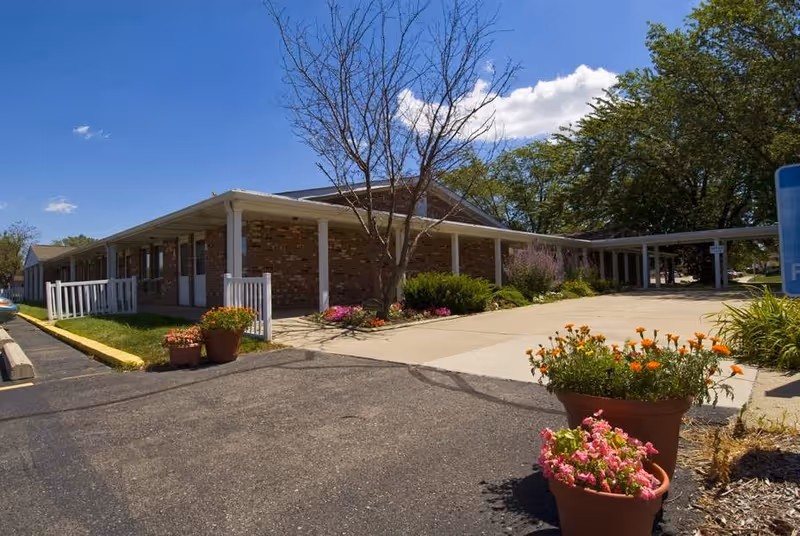 Single-story brick Maryhaven building with a covered entrance walkway, potted flowers, and a driveway under a blue sky.