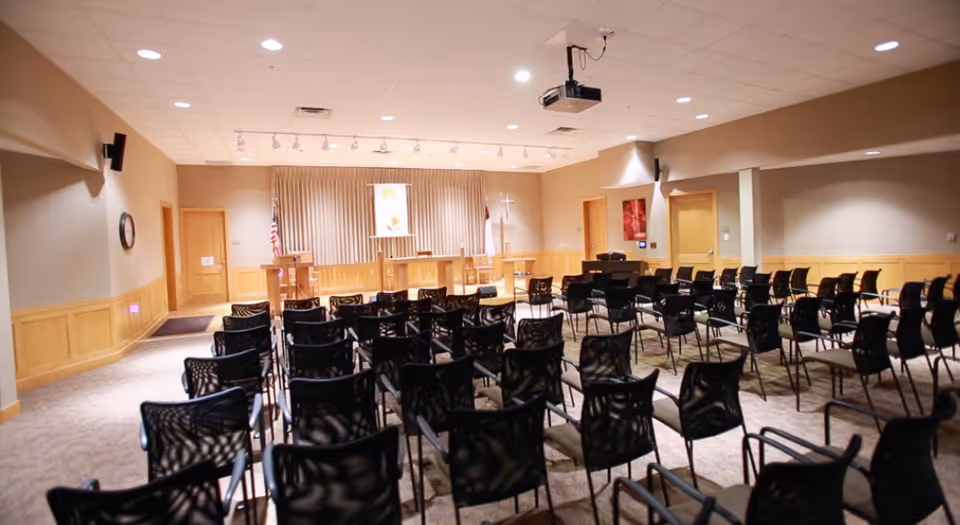 Interior view of a meeting or chapel room with rows of black chairs facing a stage area with podiums, an American flag, a Christian flag, and a cross. The room has beige walls, carpeted floor, and ceiling lights with a projector mounted on the ceiling.