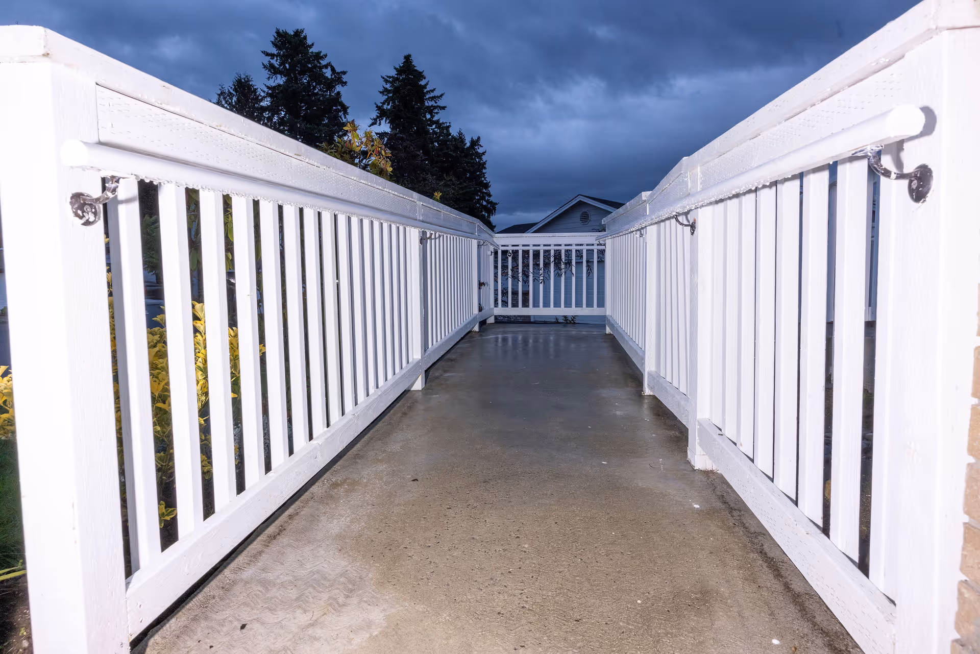 A concrete ramp with white wooden railings on both sides leading to a porch or entrance of a building, with trees and a cloudy sky in the background.