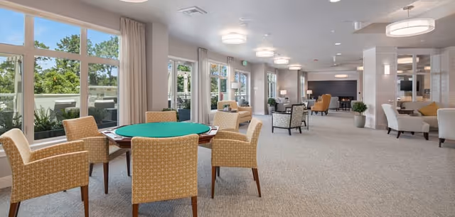 A spacious, well-lit common area with large windows showing greenery outside. The room features multiple seating arrangements including a round table with six upholstered chairs in the foreground, and several armchairs and sofas arranged around small tables further back. The decor is modern with neutral tones, carpeted floors, and ceiling lights.