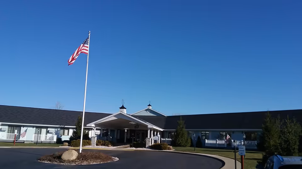 Exterior view of Arbor North Living Center building under a clear blue sky with an American flag on a flagpole in front of the entrance.