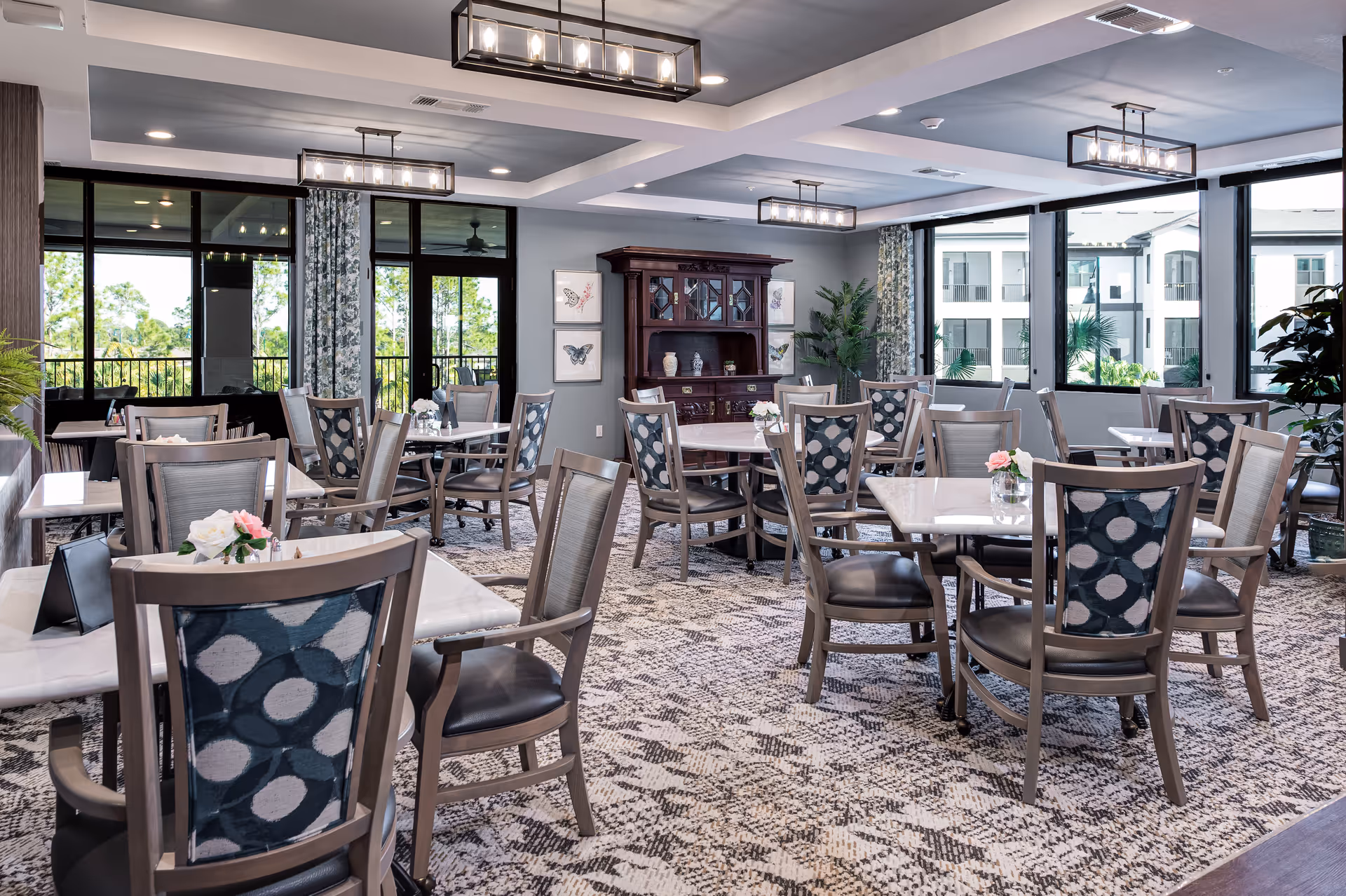 A bright and spacious dining room with multiple tables and chairs arranged neatly. The chairs have patterned backs and cushioned seats. The room features large windows with views of greenery and an adjacent building. There are decorative plants, framed butterfly artwork on the walls, and a wooden cabinet. Modern ceiling lights illuminate the space, and the floor is covered with a patterned carpet.