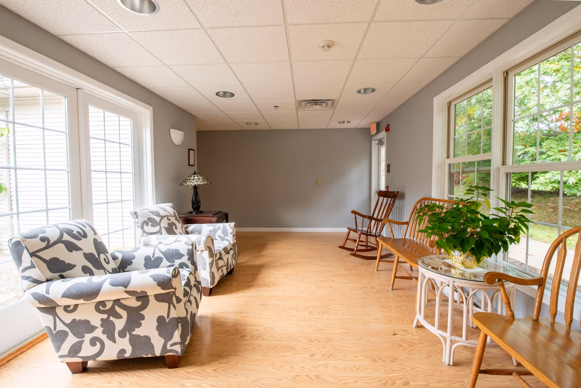 A bright sitting area with two patterned armchairs on the left, a wooden rocking chair and wooden bench on the right, a small white table with a green plant, large windows letting in natural light, and light wood flooring.