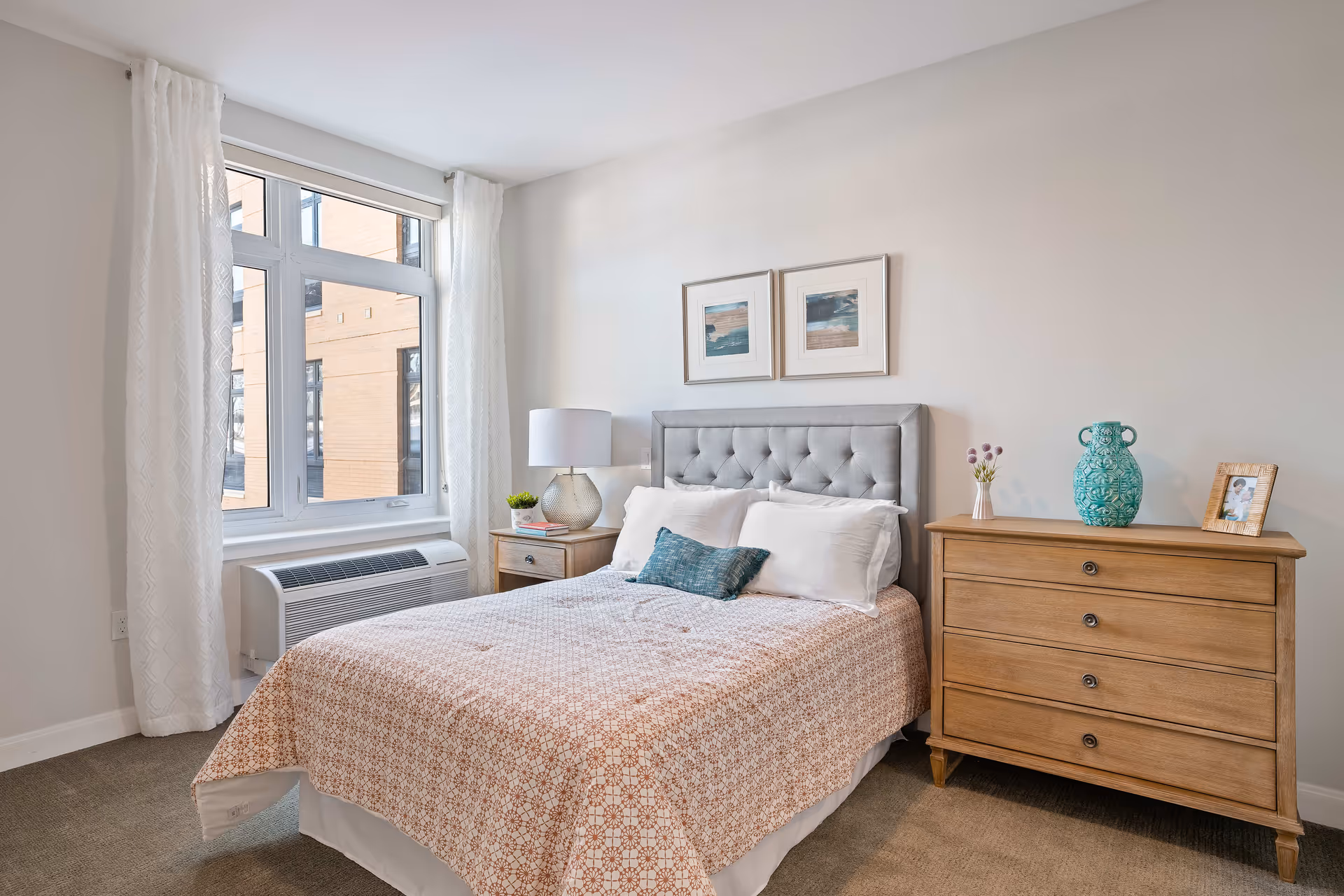A bright bedroom with a large window covered by white curtains. The room features a bed with a tufted gray headboard, white pillows, and a patterned bedspread. Next to the bed is a wooden nightstand with a lamp and a small plant. Across from the bed is a wooden dresser with a turquoise vase, a small flower arrangement, and a framed photo on top.