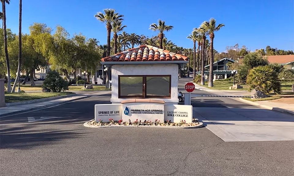 Small tiled-roof gatehouse at a landscaped entrance with palm trees and a stop sign.