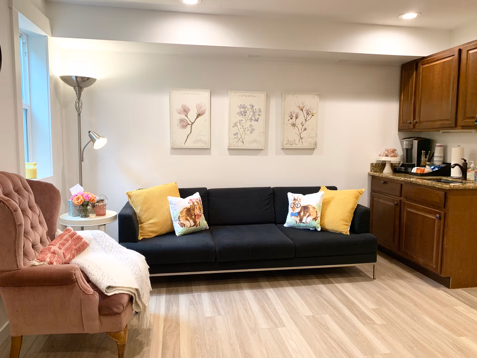 A cozy living room area with a black sofa adorned with two yellow pillows and two decorative pillows featuring animal illustrations. To the left is a pink upholstered armchair with a white throw blanket and a pink knitted pillow. A small round white side table next to the armchair holds a vase of flowers and a candle. On the wall above the sofa are three framed botanical prints. To the right, there is a wooden cabinet with a granite countertop holding a coffee maker, cups, and other kitchen items. The room has light wood flooring and white walls.