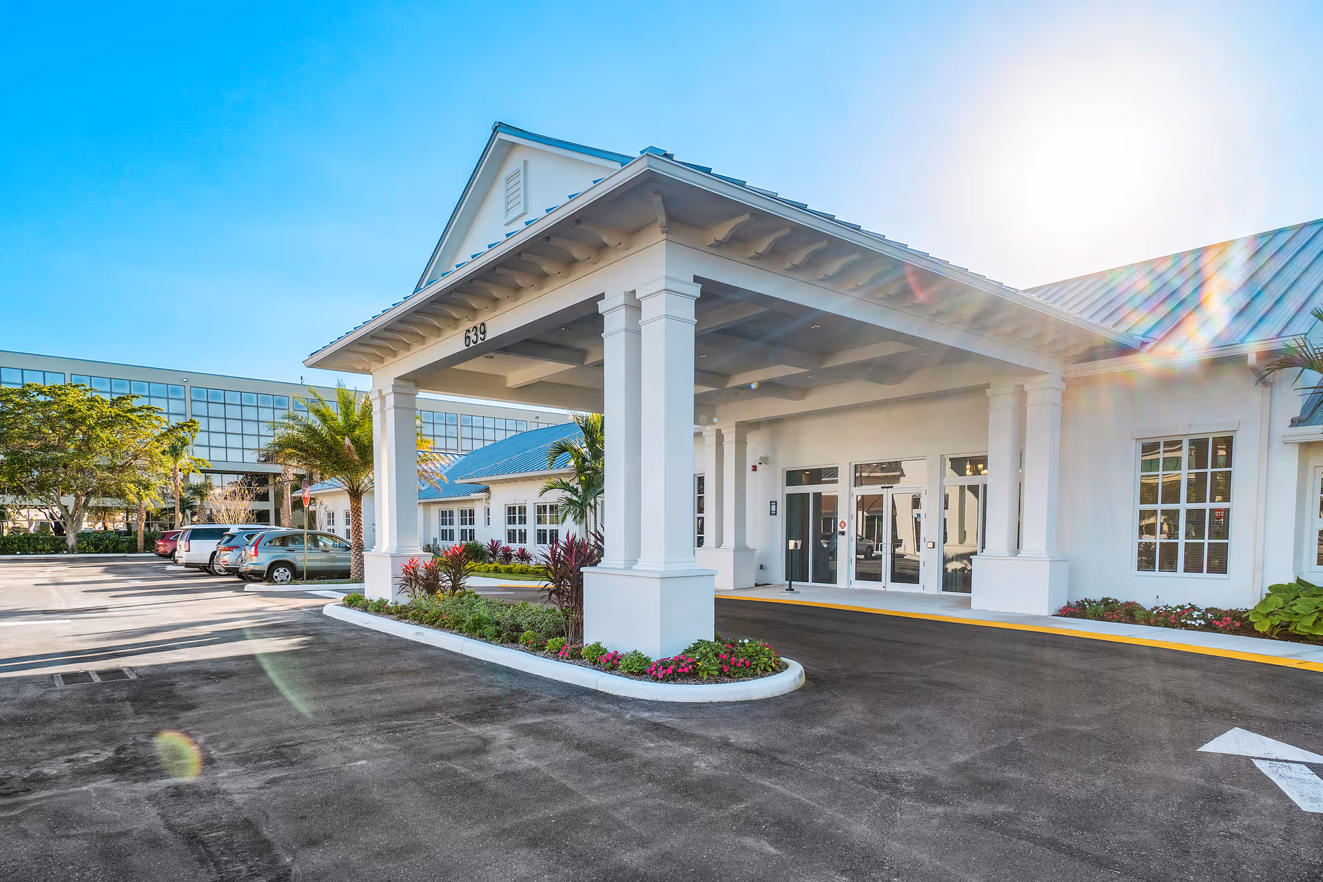 Front entrance of a white memory care facility with a covered porte-cochere, landscaping, and parked cars.