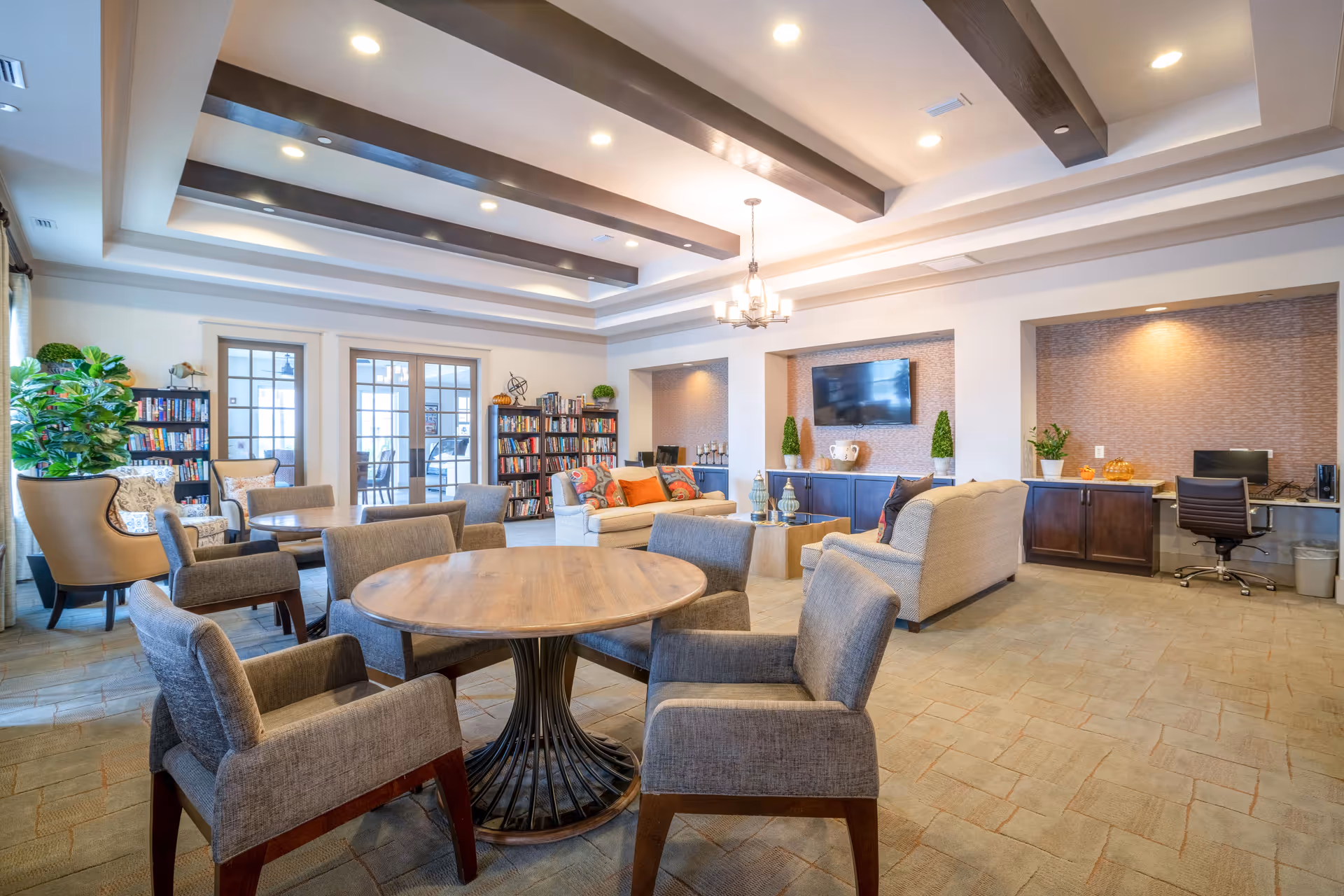 A bright and spacious living room area with multiple seating options including armchairs and sofas arranged around a coffee table. There are two round wooden tables with cushioned chairs. The room features a ceiling with wooden beams and recessed lighting. A flat-screen TV is mounted on the wall above a cabinet with decorative plants and pumpkins. Bookshelves filled with books are visible near glass double doors, and a small desk with a computer is set into a recessed nook.
