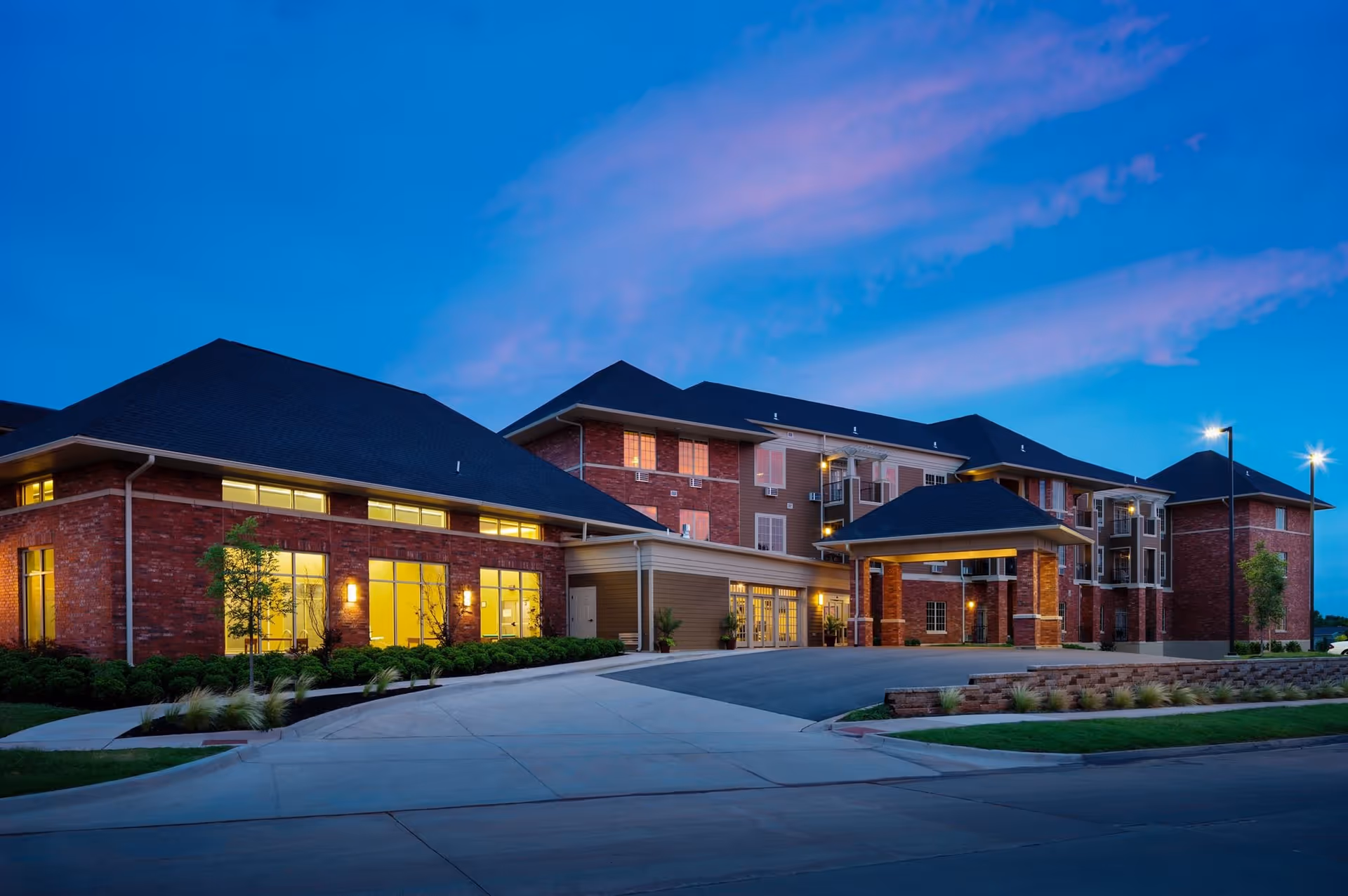 Front exterior of a large brick senior living building at dusk with lit windows and a covered entrance.