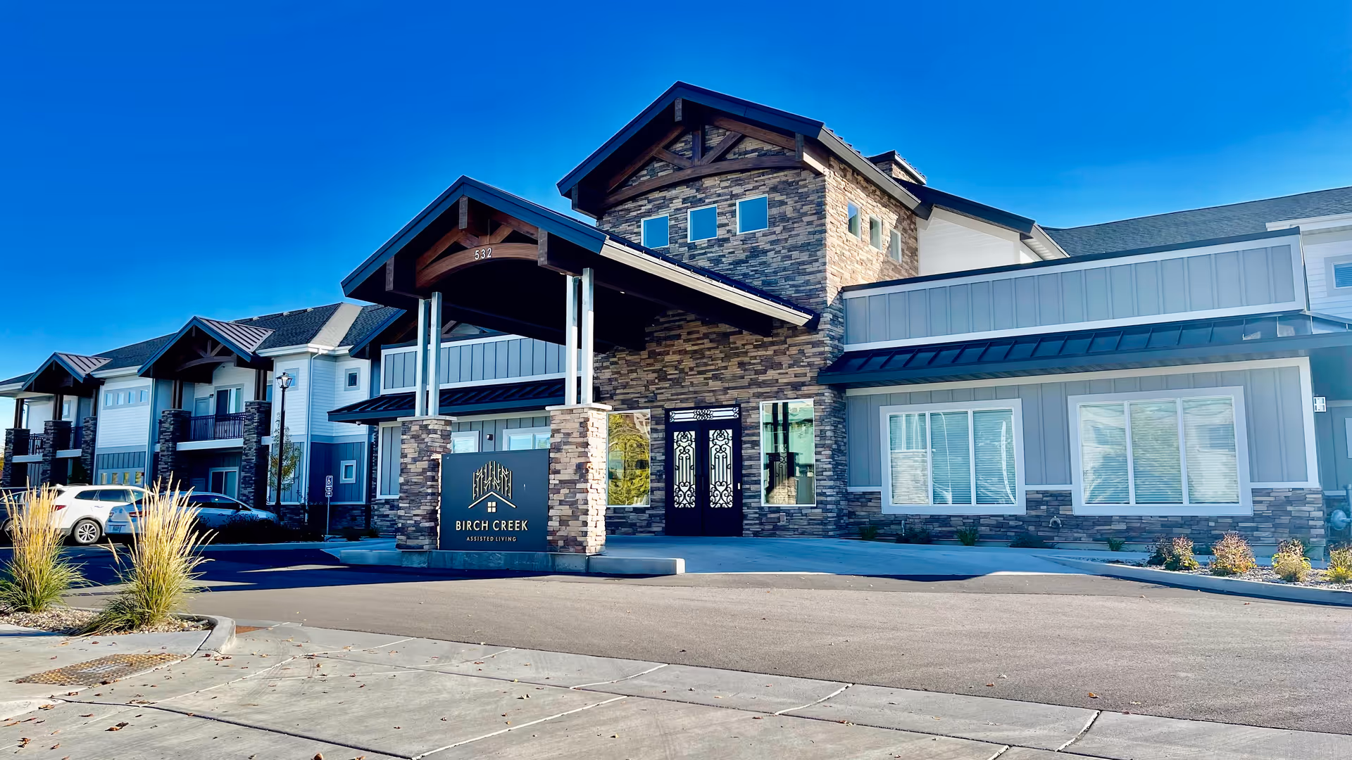 Front entrance of Birch Creek Assisted Living building with a covered porte-cochère, stone facade, and clear blue sky.