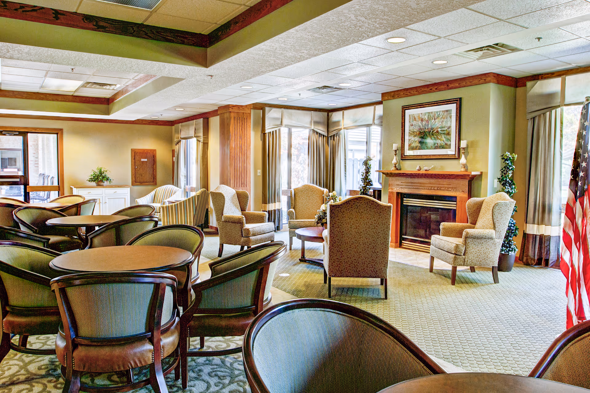 A cozy senior living facility common area with multiple round tables and chairs in the foreground. In the background, there are several upholstered armchairs arranged around a wooden fireplace with a framed painting above it. Large windows with curtains allow natural light to fill the room, and an American flag stands near the window on the right side.