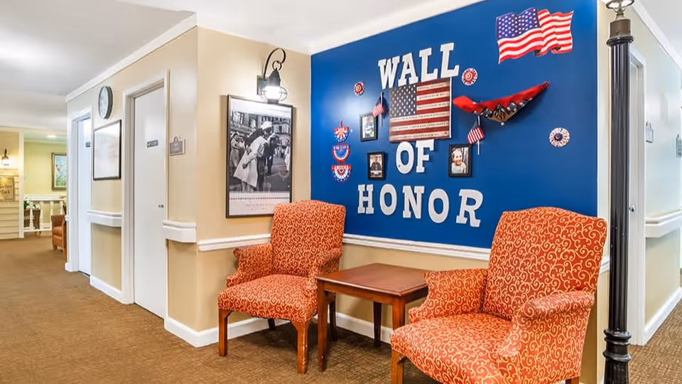 A hallway area in a senior living facility with two patterned orange armchairs and a small wooden table between them. On the wall behind the chairs is a blue display titled 'Wall of Honor' decorated with American flags, photos, and patriotic items. The hallway has beige walls, carpeted floor, and a black lamp post style light fixture on the right side.