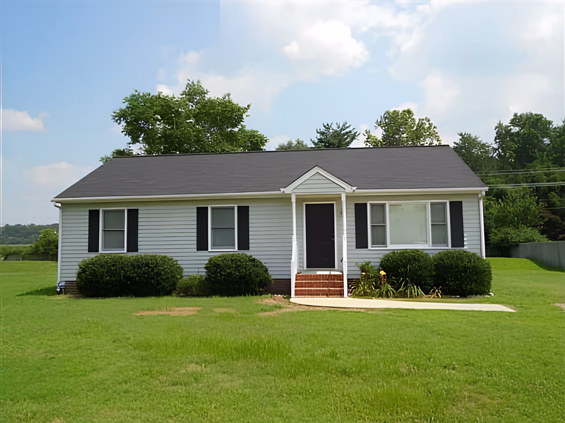 Single-story white house with black shutters and a black roof, surrounded by green grass and bushes, under a partly cloudy blue sky.