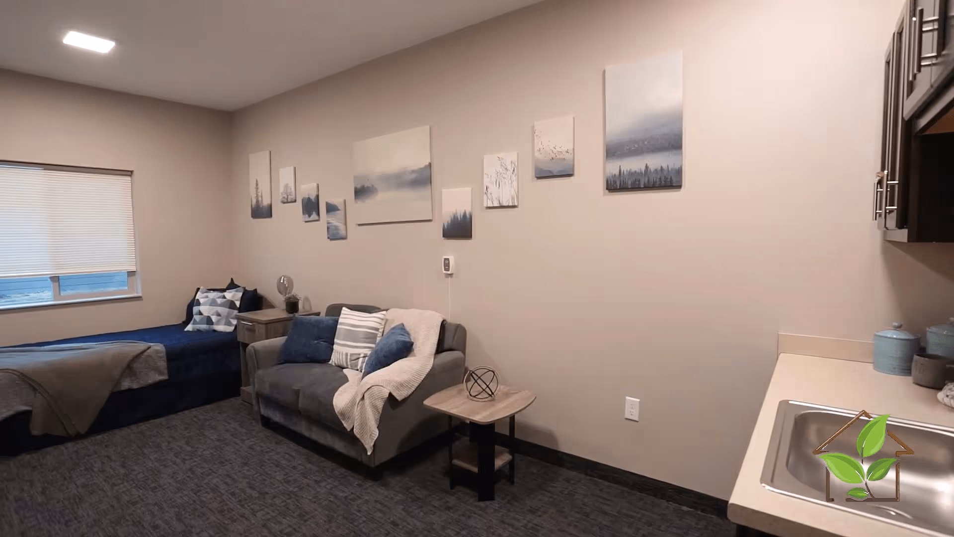 A cozy room with a single bed covered in blue bedding and a gray blanket, a small gray loveseat with blue and striped pillows and a beige throw, a wooden side table with a decorative metal sphere, and a series of landscape paintings on the beige wall. There is a window with a closed blind and a small kitchenette area with a sink and countertop on the right side.