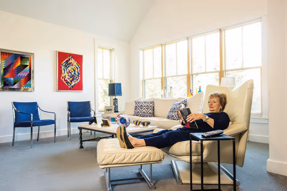 An elderly woman is reclining on a beige leather lounge chair with a matching ottoman in a bright living room. She is reading a book and has a remote control on a small black side table next to her. The room features a beige sofa with patterned cushions, two blue chairs with metal frames, a coffee table with decorative items, and two colorful geometric paintings on the wall. Large windows with blinds allow natural light to fill the space.
