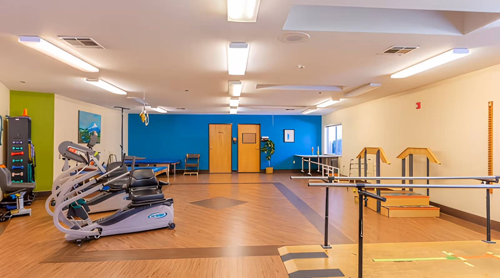A spacious rehabilitation room with exercise equipment including stationary bikes and parallel bars for physical therapy. The room has a wooden floor, bright overhead lighting, a blue accent wall with two wooden doors, and a green wall section with exercise accessories. There are also therapy tables and chairs along the walls.