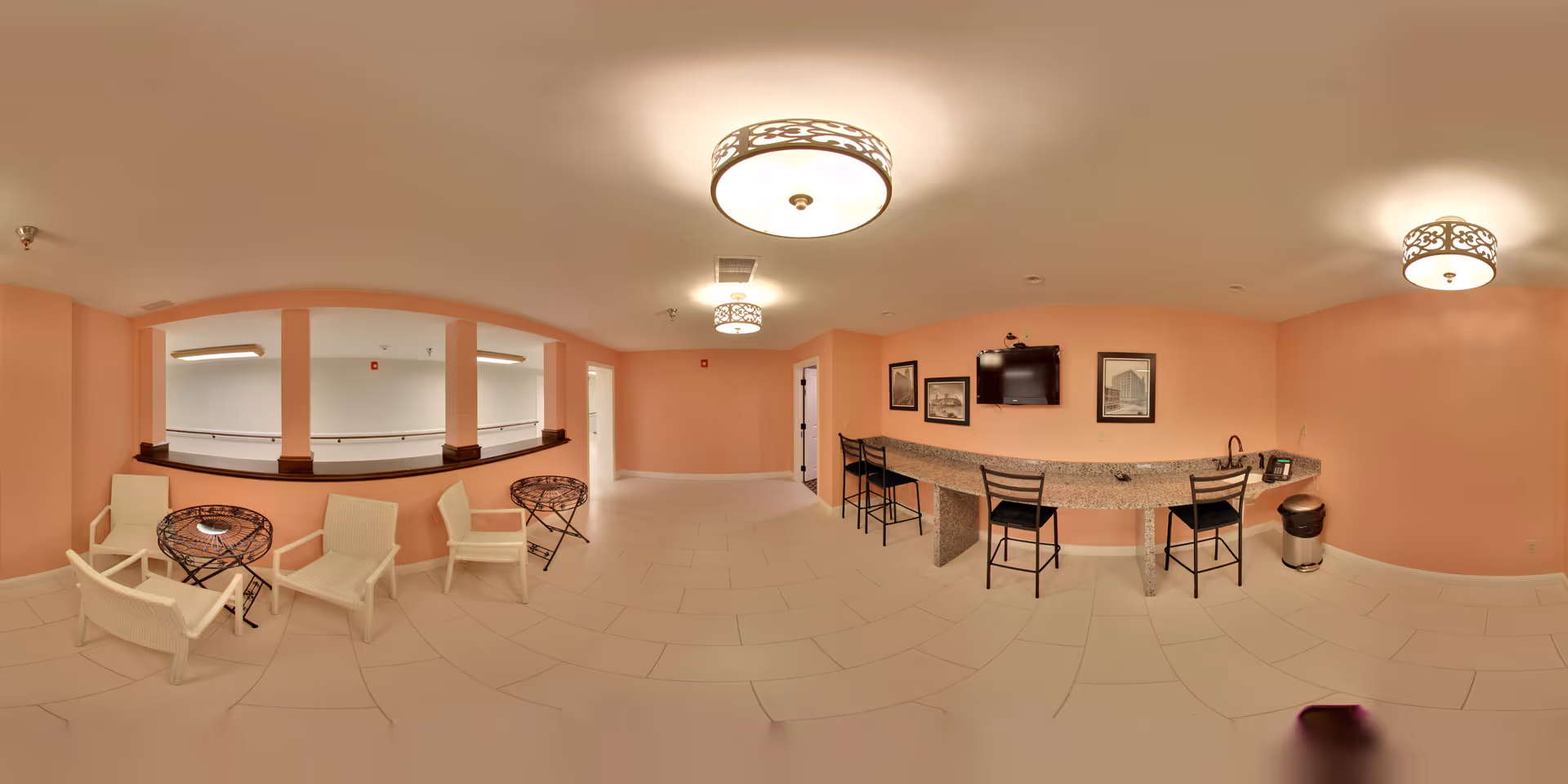 A spacious interior common area with peach-colored walls and tiled floor. The room features a long granite countertop with four black chairs, a wall-mounted TV, and framed pictures on the wall. On the left side, there are four white chairs and two small round tables near a half-wall with windows looking into another room. Ceiling lights provide warm illumination.