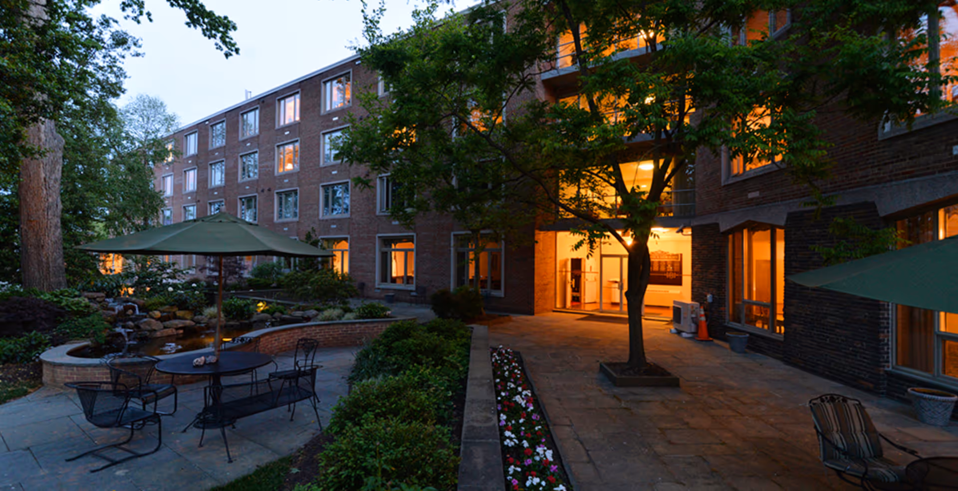 Outdoor patio area at dusk with metal tables and chairs under green umbrellas, surrounded by greenery and flowers, adjacent to a multi-story brick building with lit windows.