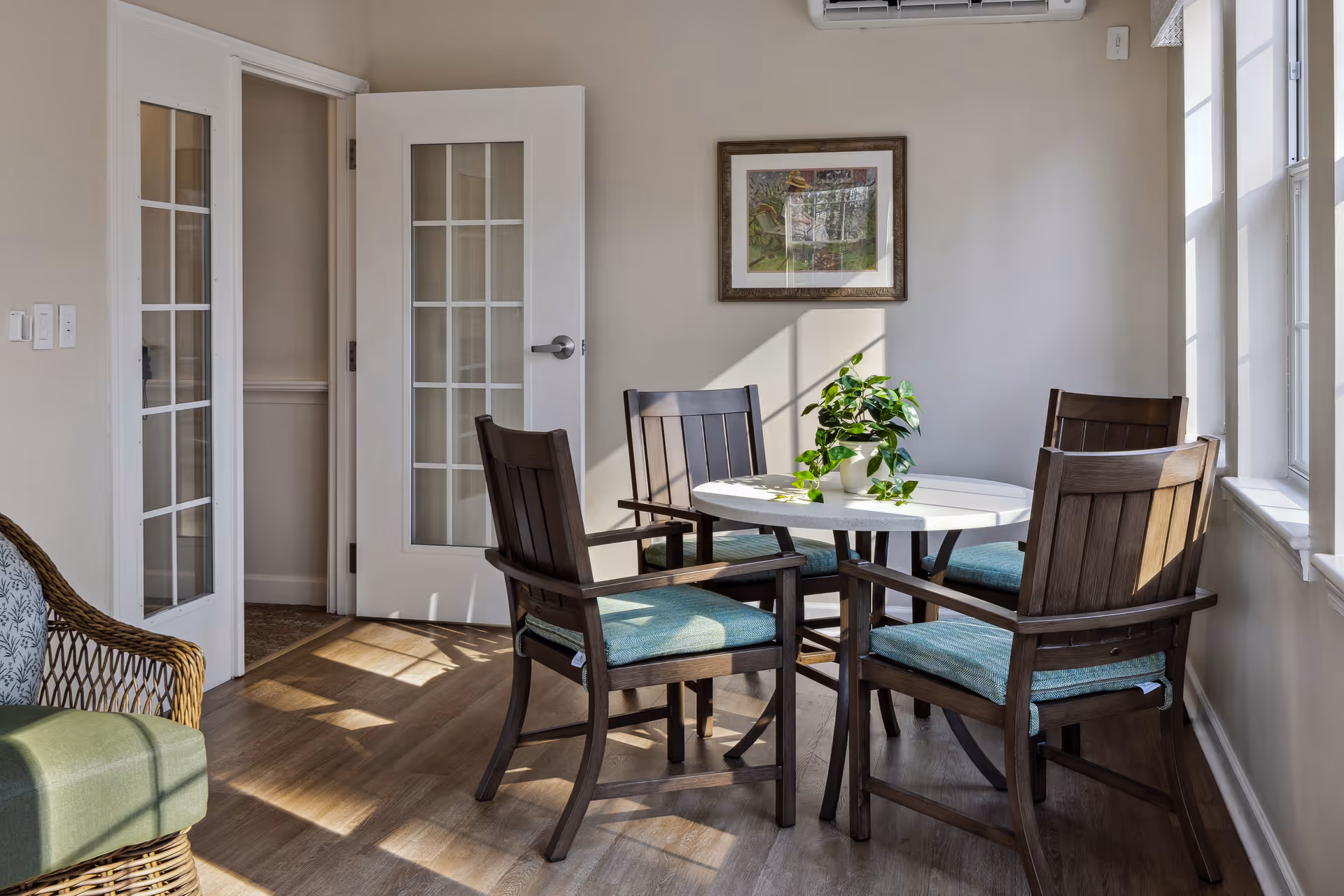Sunlit dining area with a round table, four wooden chairs with teal cushions, a potted plant centerpiece, and French doors.