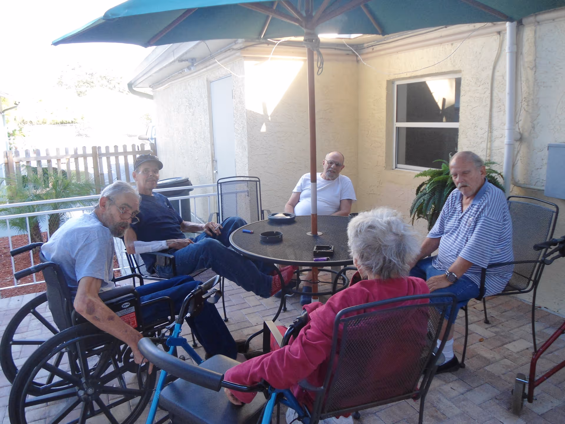 Five elderly individuals sitting around a round outdoor table with a large umbrella on a patio area. Two of the individuals are in wheelchairs, and the group appears to be engaged in conversation. The setting is outside a building with a window and a door visible, along with some plants and a fence in the background.