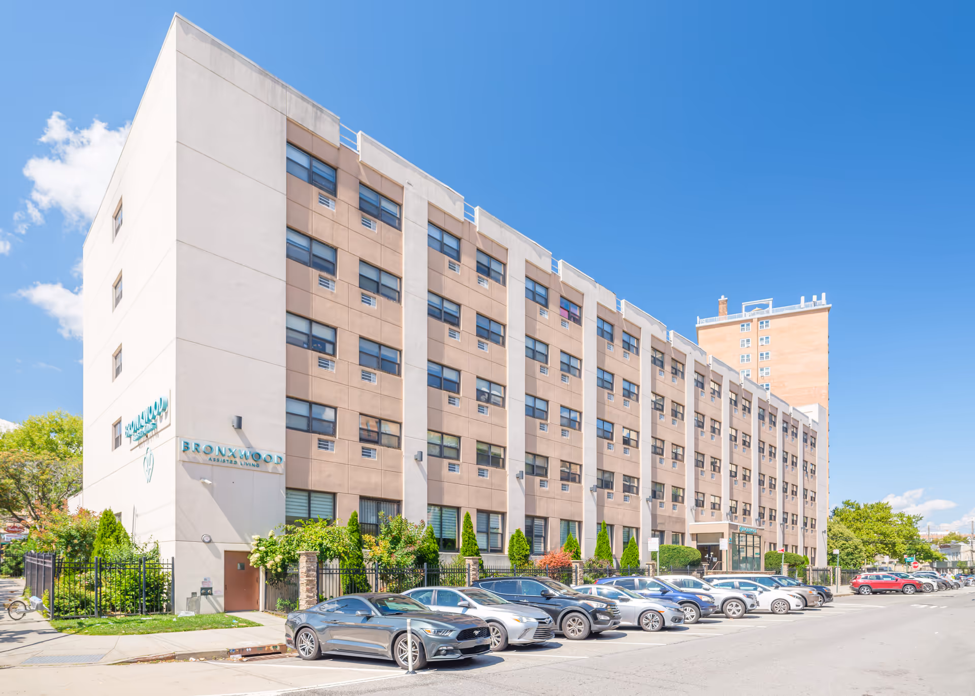 Exterior view of Bronxwood Assisted Living facility, a multi-story building with numerous windows and a parking lot in front filled with cars under a clear blue sky.