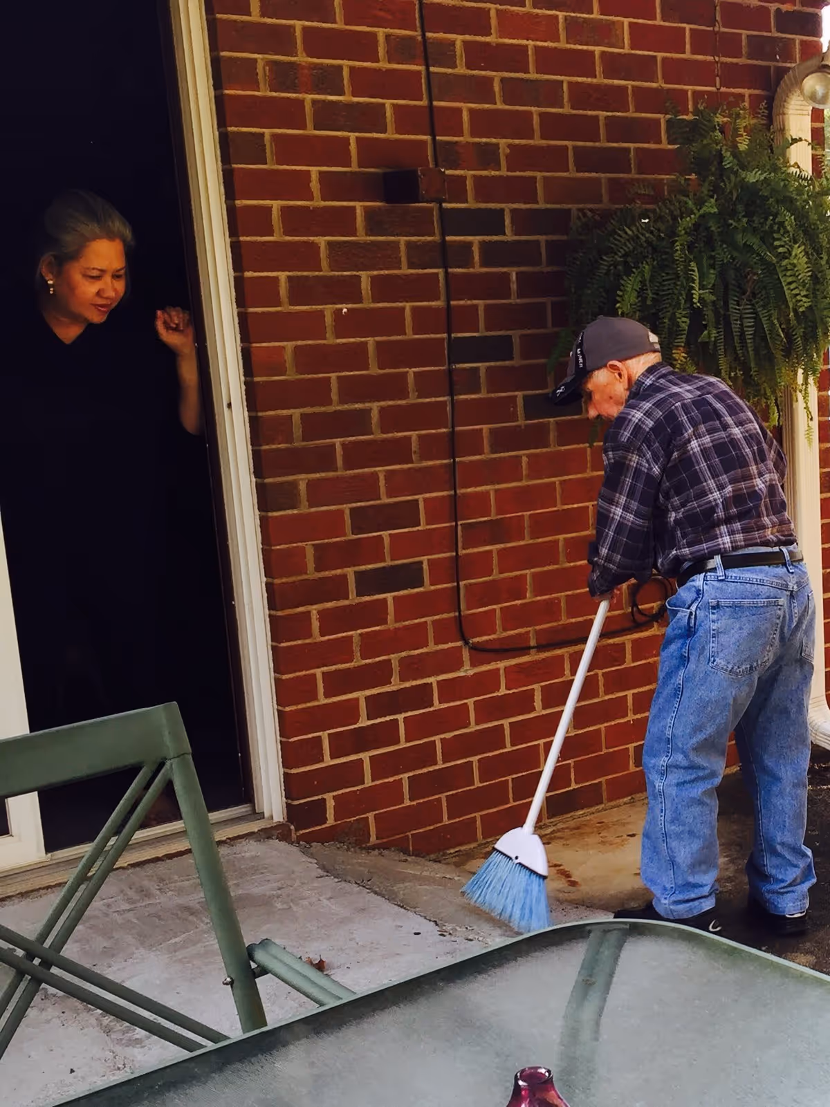 An elderly man wearing a plaid shirt, jeans, and a cap is sweeping the concrete floor outside a brick building. A woman with gray hair is standing in the doorway watching him. There is a green metal chair and a glass table with a small purple vase in the foreground, and a hanging green fern plant on the wall.