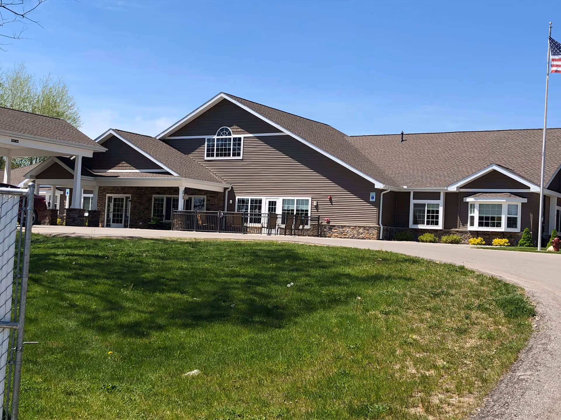 Front exterior of a single-story senior living building with gabled roofs, a driveway, and a grassy lawn with an American flag.