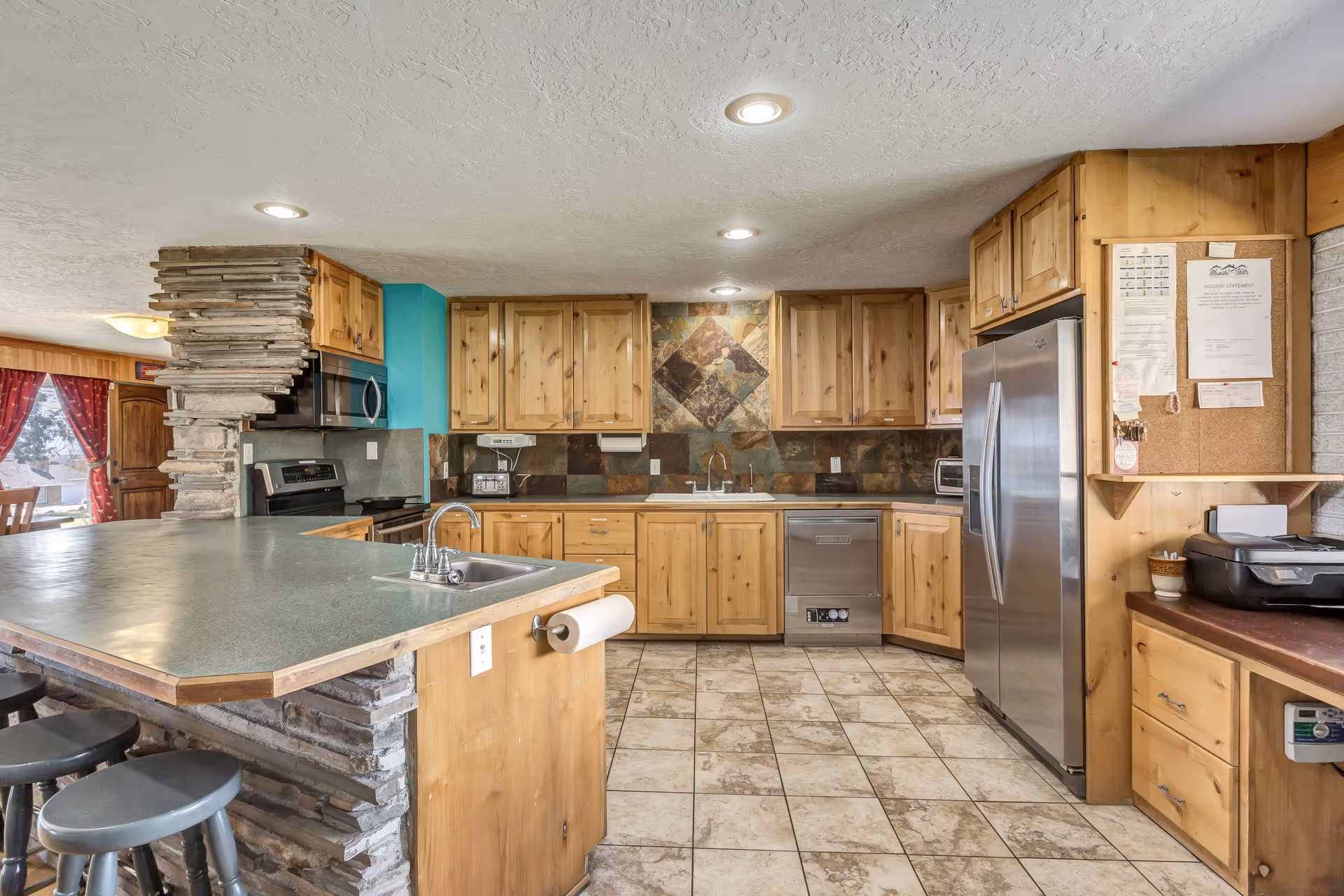 A spacious kitchen with wooden cabinets, a stone accent wall, and a large countertop with a built-in sink. The kitchen features stainless steel appliances including a refrigerator, microwave, and dishwasher. There are bar stools along the counter and a bulletin board with papers pinned on the right side.