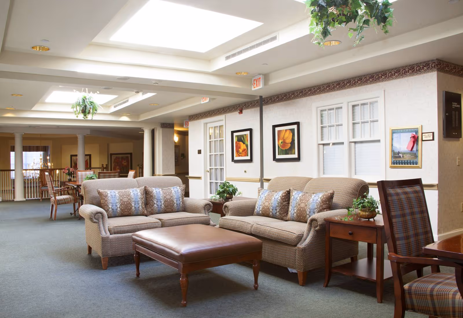 A bright and spacious common area in a senior living facility featuring two beige sofas with patterned cushions, a large brown ottoman, wooden side tables with plants, and framed floral artwork on the walls. The room has skylights, carpeted floors, and a dining area with wooden chairs and tables in the background.