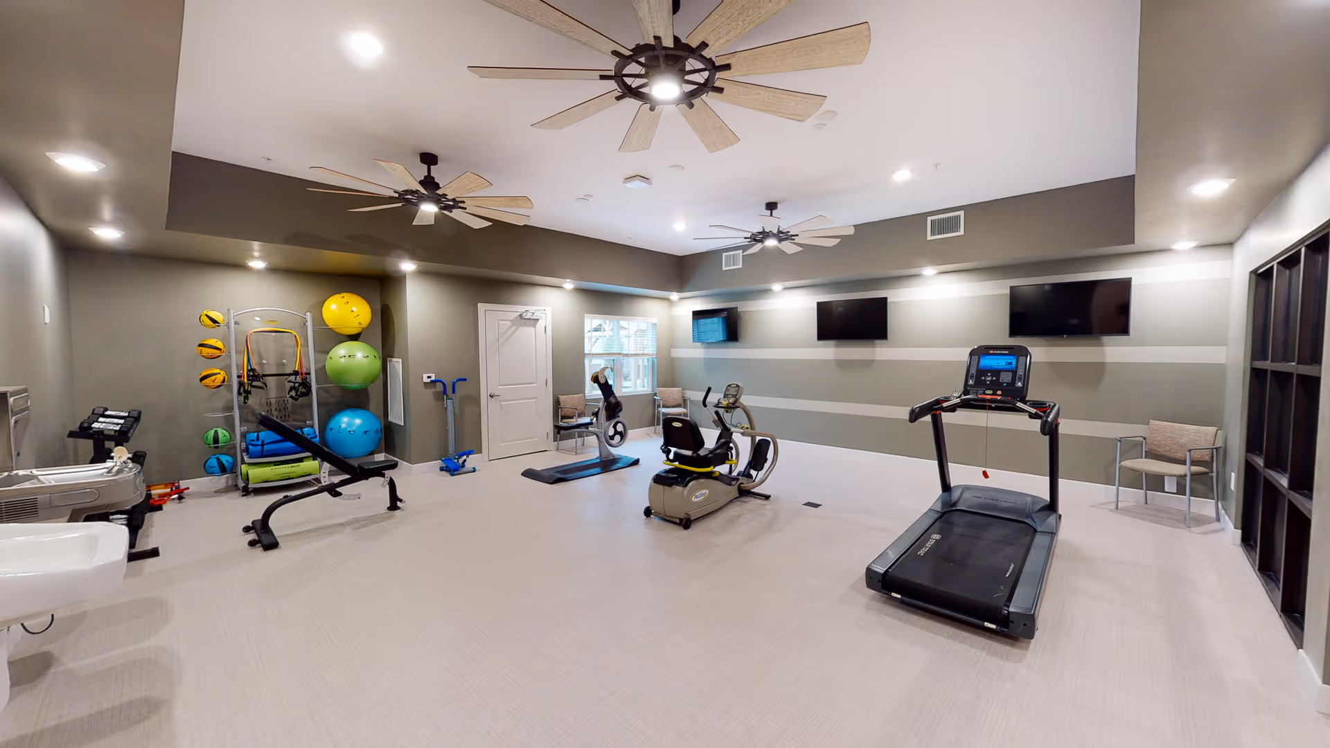 A spacious fitness room with exercise equipment including a treadmill, recumbent bike, elliptical machine, and a weight bench. The room has light-colored flooring, gray walls with horizontal stripes, three ceiling fans, and several colorful exercise balls and resistance bands stored on a rack. There are also two chairs near a window and two wall-mounted televisions.