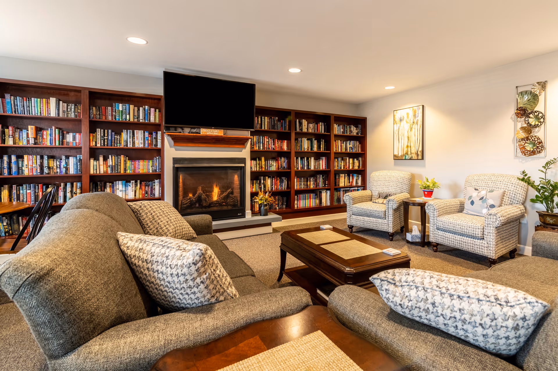 Cozy living room with a gray sofa and patterned pillows, two armchairs, a wooden coffee table, built-in bookshelves filled with books, a fireplace with a fire burning, and a flat-screen TV mounted above the fireplace. The room is warmly lit with recessed ceiling lights and decorated with wall art and plants.