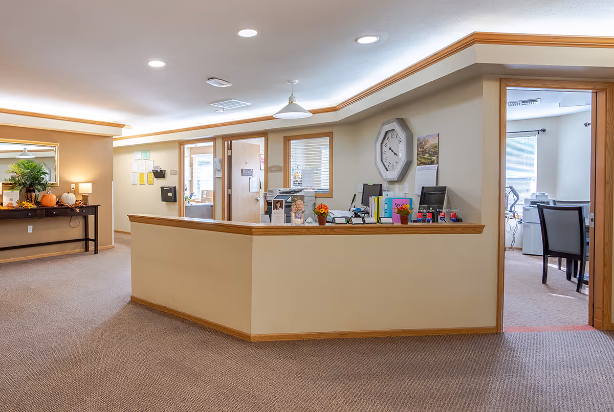 Reception area in an assisted living facility with a beige carpet, a wooden reception desk, and a wall clock. There is a table with fall decorations including pumpkins and a plant on the left side. Two open doorways lead to office spaces with desks and chairs.