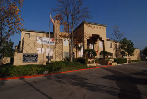 Front exterior of a two-story beige building with a recessed arched entrance, American flag, trees, and landscaped frontage by a parking area.