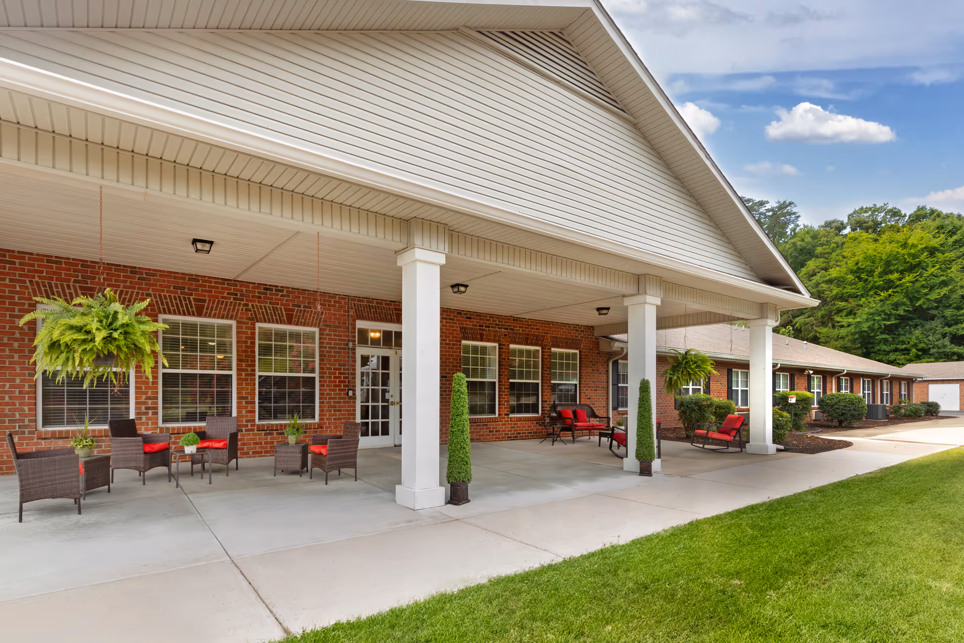 Covered front entrance of a brick senior living facility with patio seating, hanging plants, and white columns.