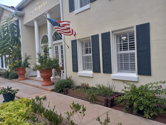Exterior view of a beige building with white columns at the entrance, two windows with dark blue shutters, and an American flag mounted near the door. There are large potted plants and a garden with various green plants and flowers along the sidewalk. The building has the name 'St. Angelus' displayed above the entrance.