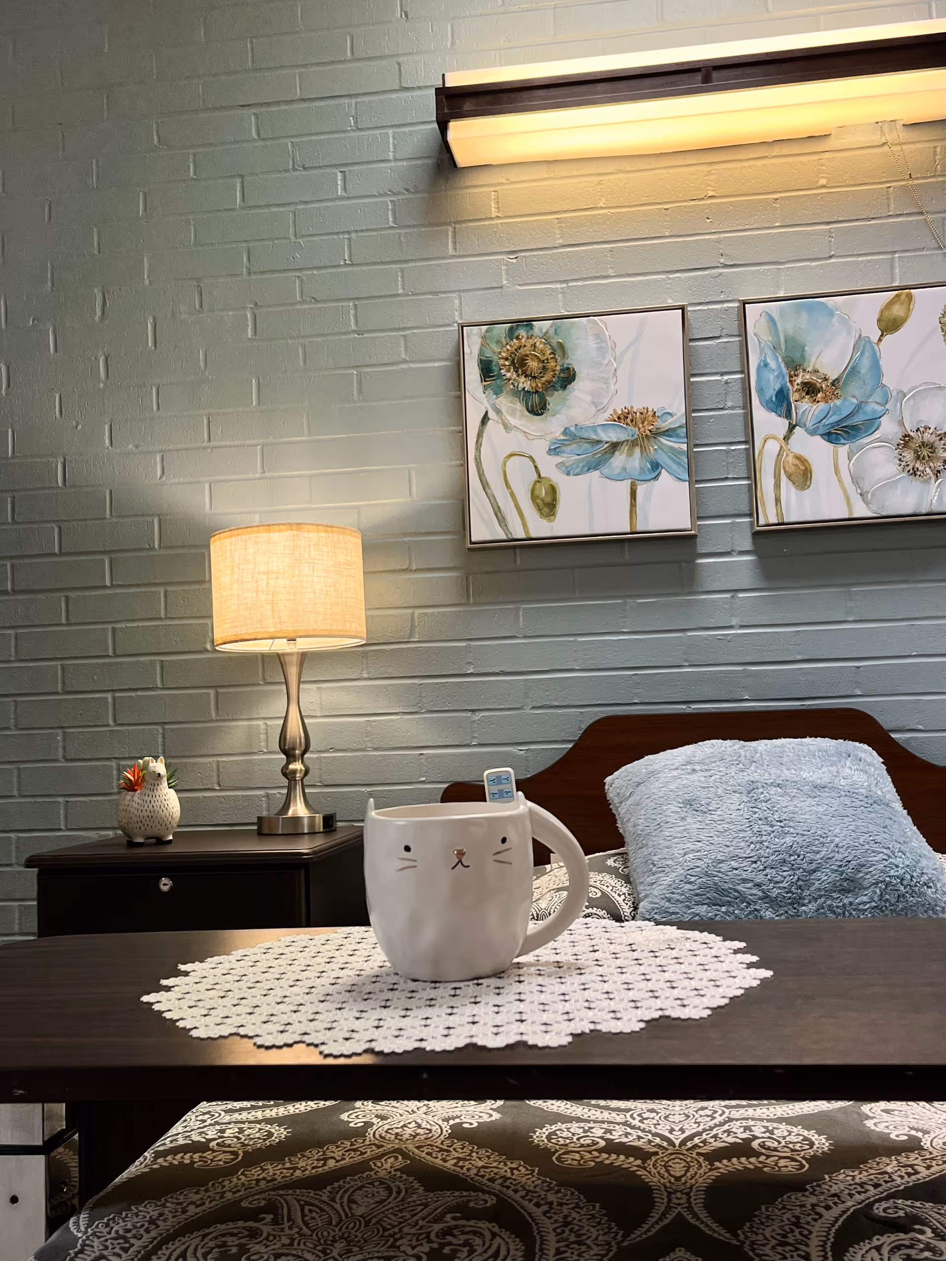 A cozy bedroom scene with a wooden bed featuring a blue fluffy pillow and patterned bedding. On a dark wooden table in the foreground, there is a white cat-shaped mug placed on a white crocheted doily. Behind the bed, a nightstand holds a lamp with a beige shade and a small decorative owl. The wall is painted light gray with a brick texture and decorated with two framed floral paintings in shades of blue and green. A long horizontal light fixture is mounted above the paintings.