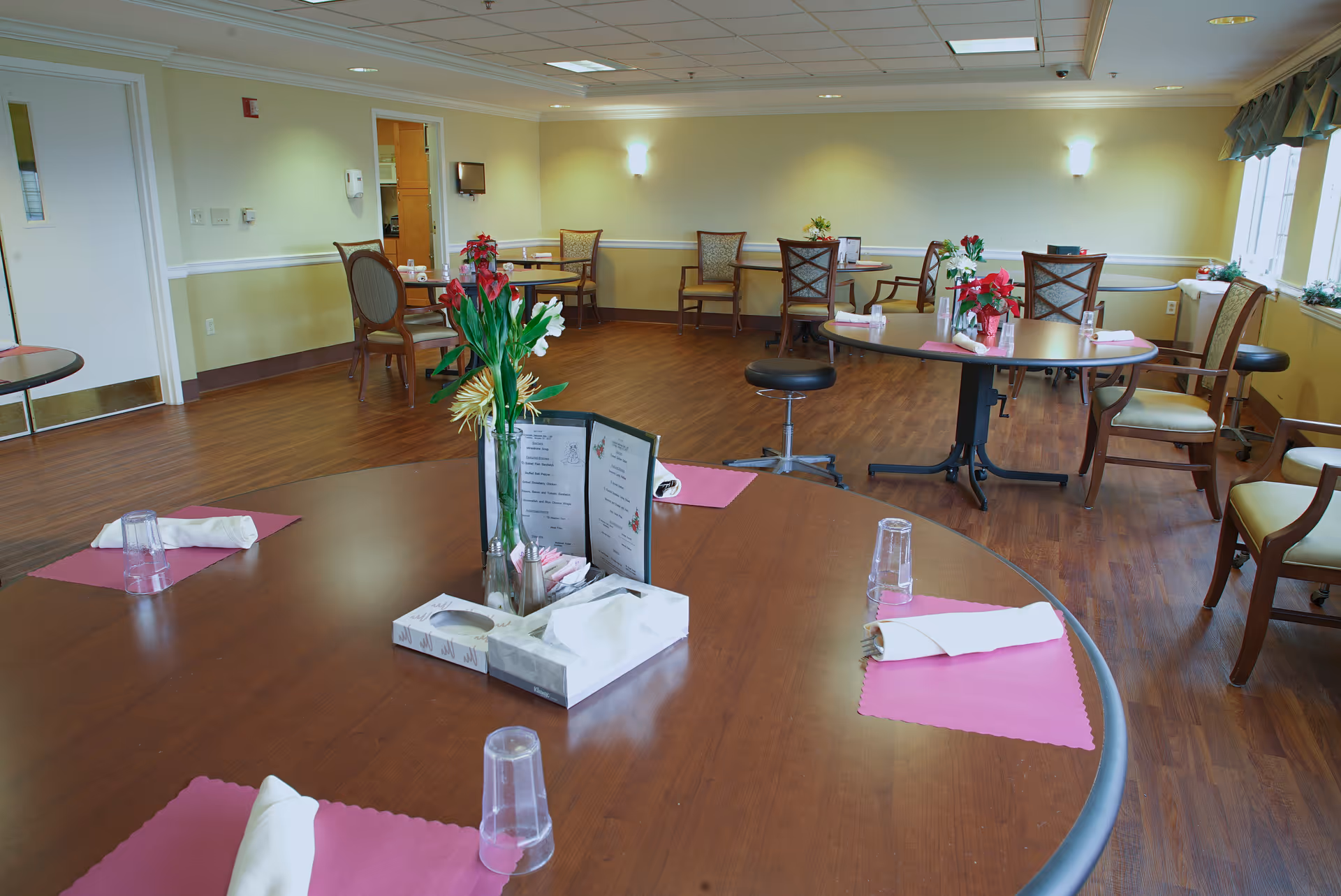 A dining room in a senior living facility with several round tables set with pink placemats, rolled white napkins, clear plastic cups, and floral centerpieces. The room has wooden flooring, beige walls, and multiple chairs arranged around the tables. There is a door leading to another room and windows on the right side letting in natural light.