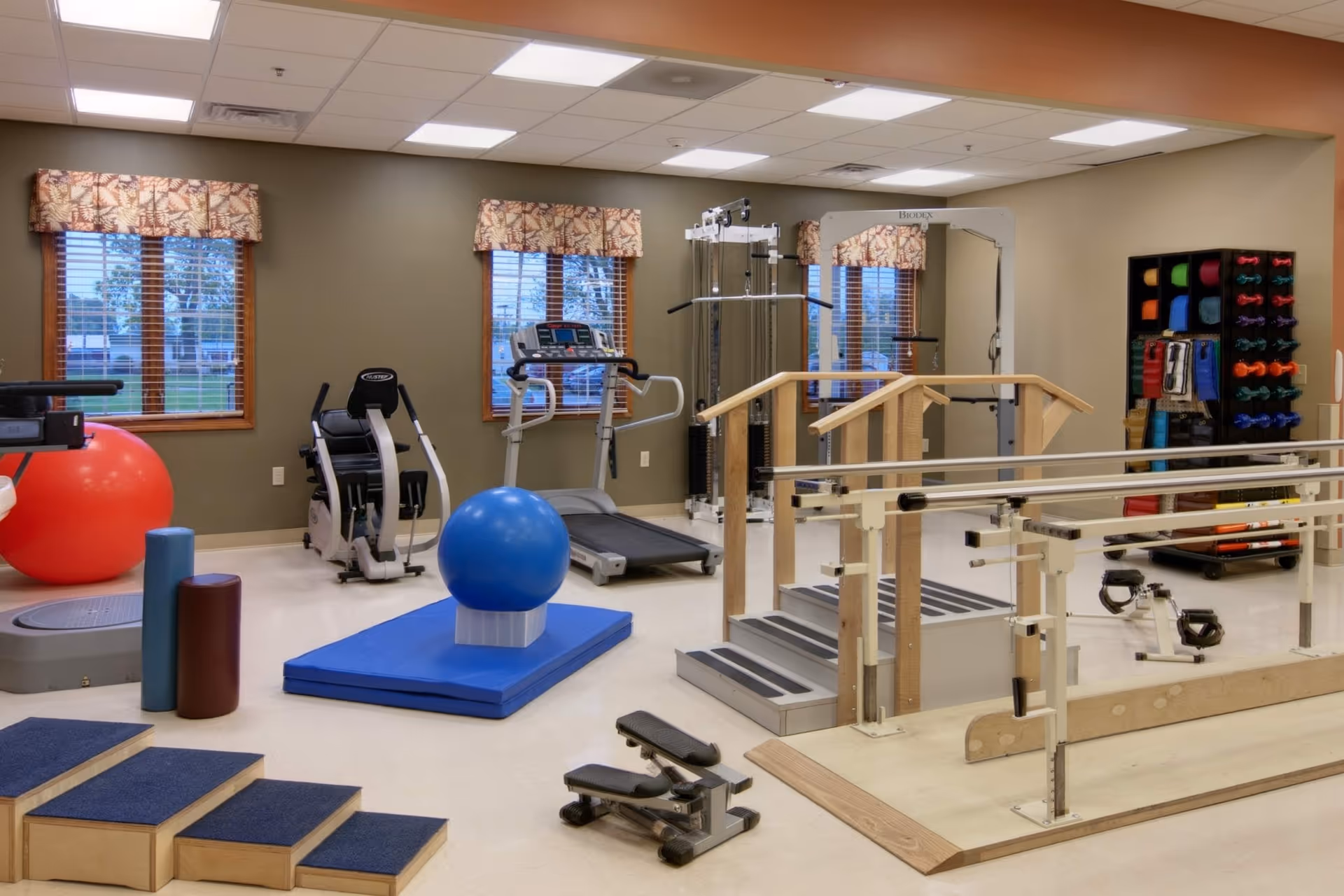 Therapy and fitness room with parallel bars, treadmill, exercise machines, balance balls, and assorted rehab equipment.