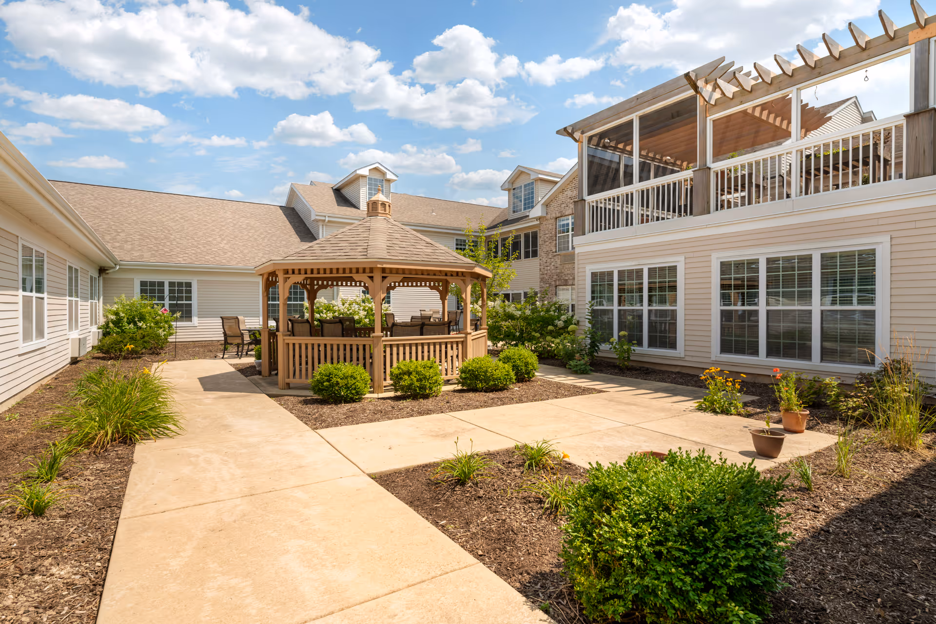 Sunny courtyard featuring a wooden gazebo, paved walkways, landscaping, and an adjacent multi-story residential building.