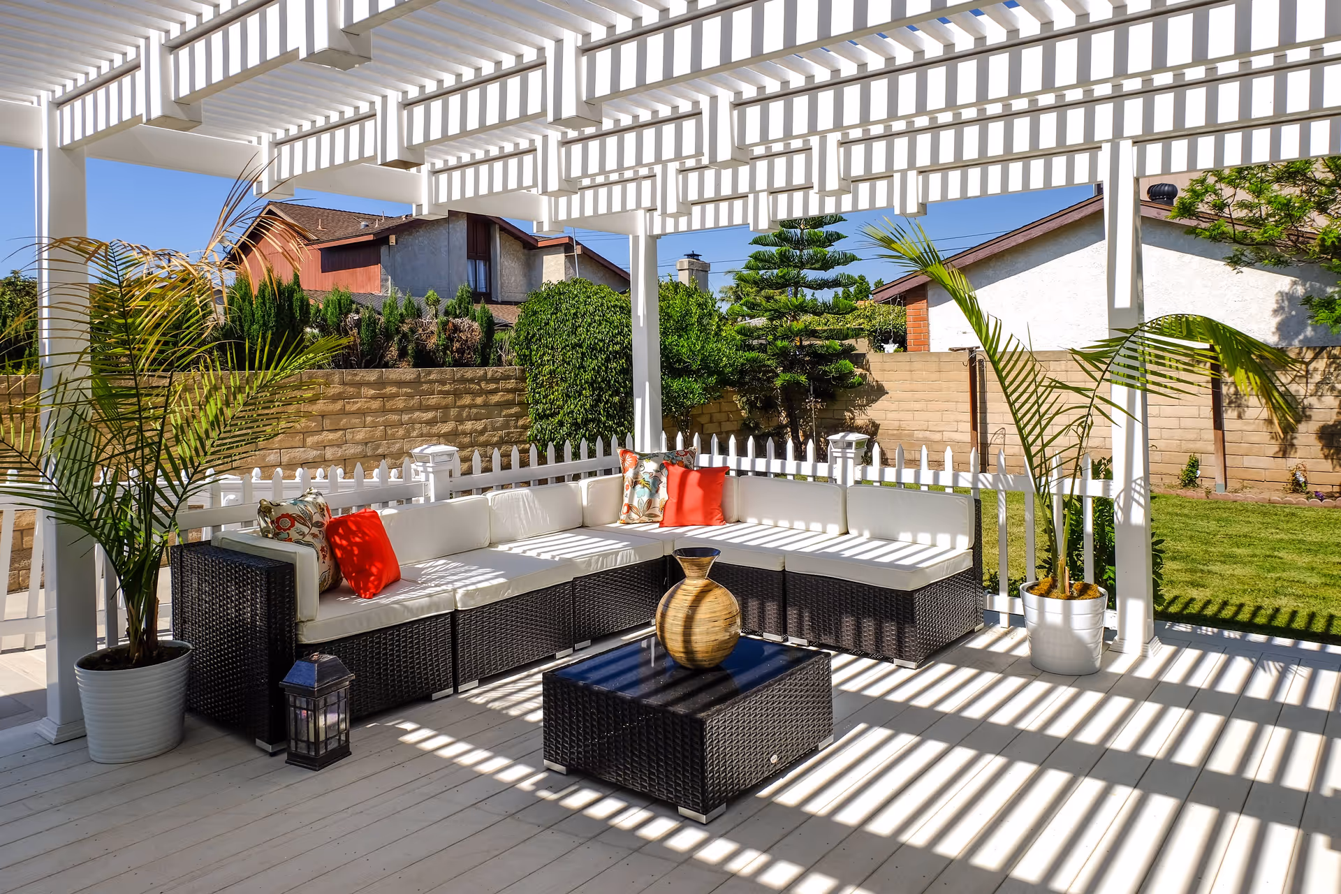 Outdoor patio area with a white pergola casting striped shadows on a wicker sectional sofa with white cushions and colorful pillows. There is a matching wicker coffee table with a decorative vase on it, two potted palm plants, a small lantern, and a white picket fence enclosing the space. In the background, there are green bushes, a brick wall, and neighboring houses under a clear blue sky.