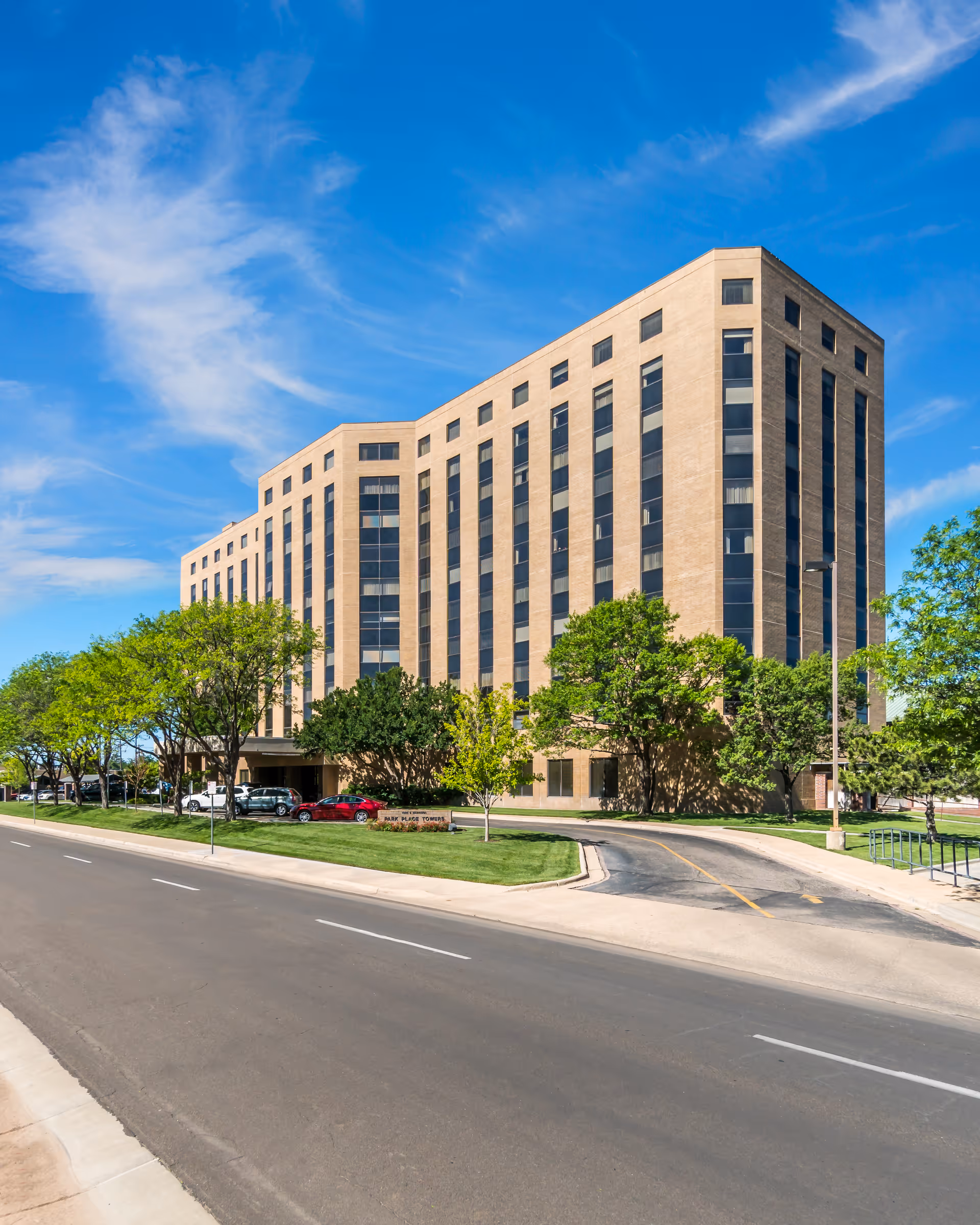 Exterior view of a multi-story beige brick building with many windows, surrounded by green trees and a well-maintained lawn under a blue sky with some clouds. Several cars are parked near the entrance along a curved driveway.